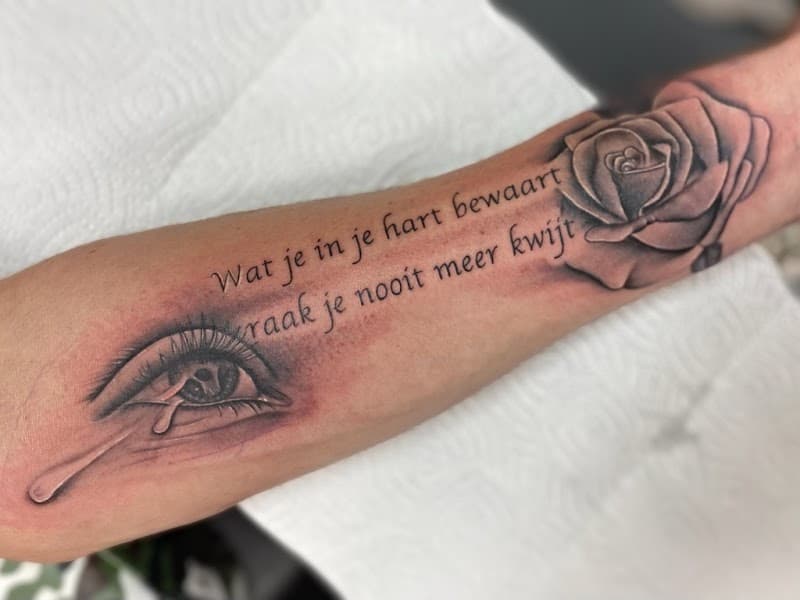 Zwart-wit tattoo van een huilend oog, roos en Nederlandse tekst op de onderarm.
