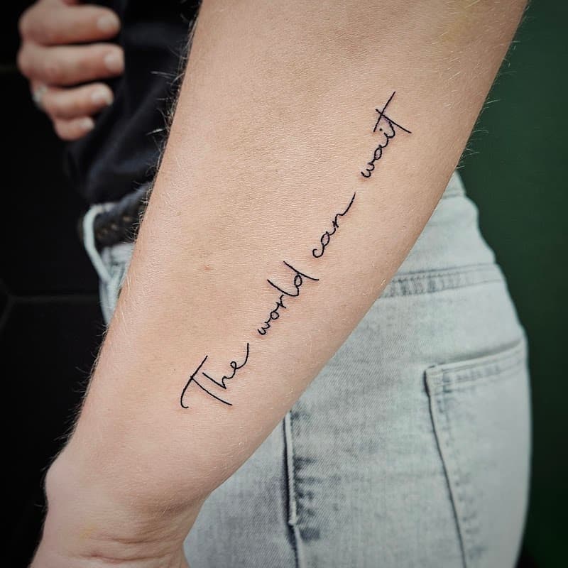 Fineline tekst tattoo 'The world can wait' op onderarm.