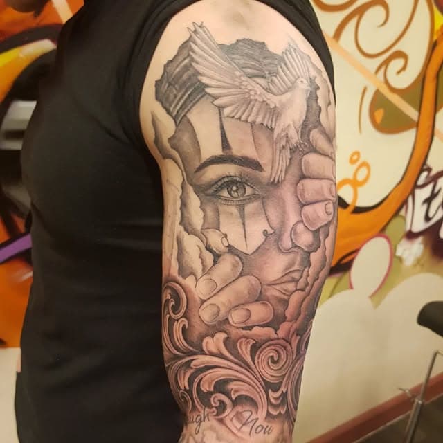 Zwart-wit tattoo van een gezicht met duif, handen en filigraan op de bovenarm.