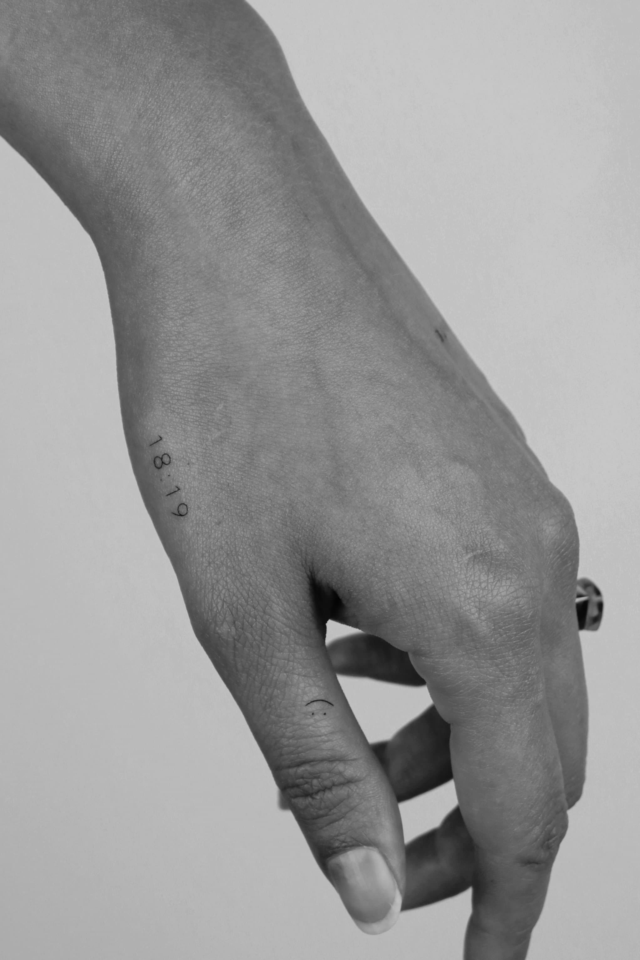Minimalistische '18:19' en smiley tattoo op hand en duim.
