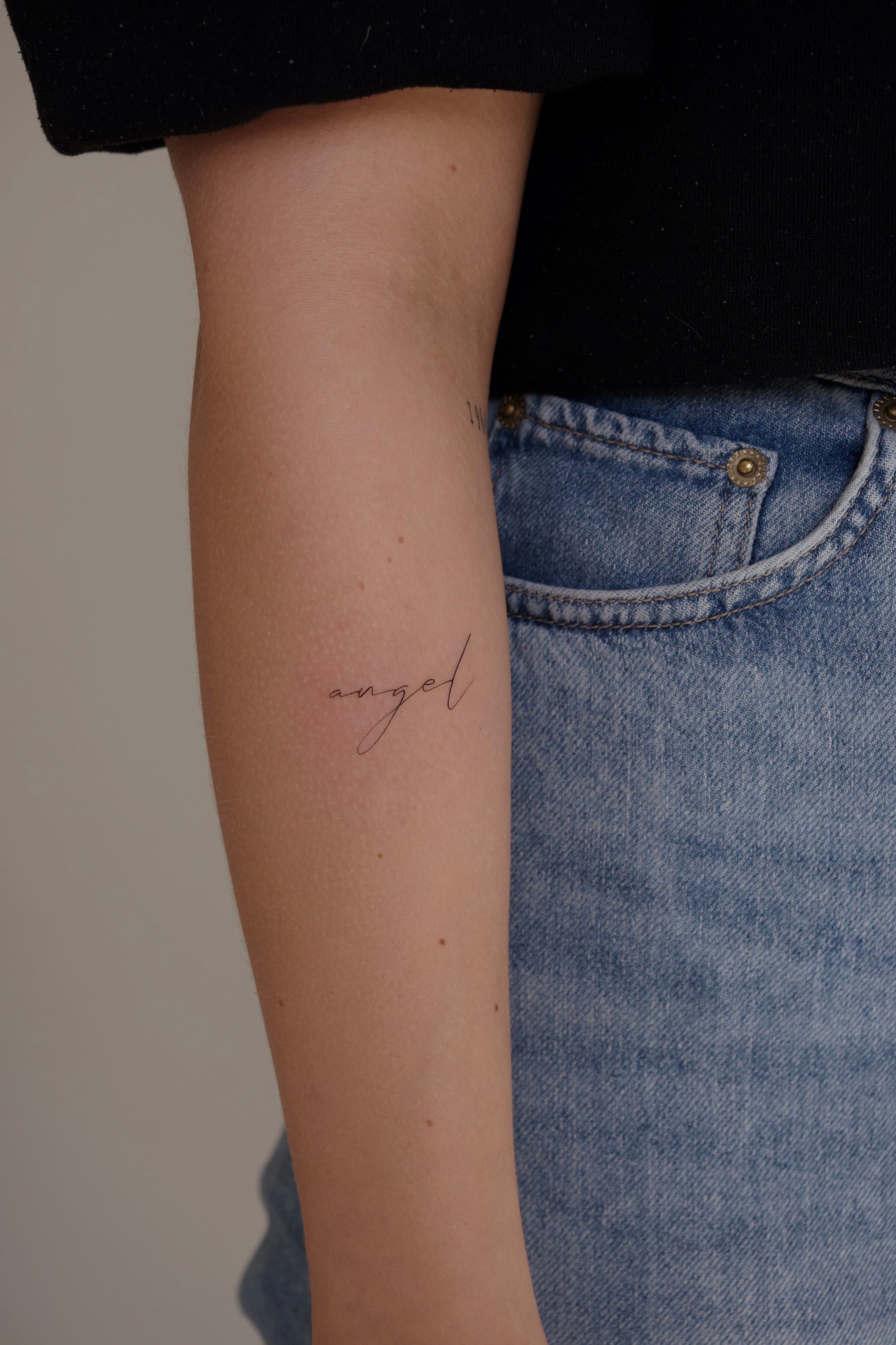 Fijne 'angel' tekst tattoo in sierlijk handschrift op de onderarm.