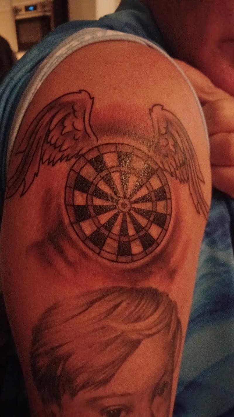 Zwart-wit tattoo van een dartbord met vleugels en een portret op de bovenarm.