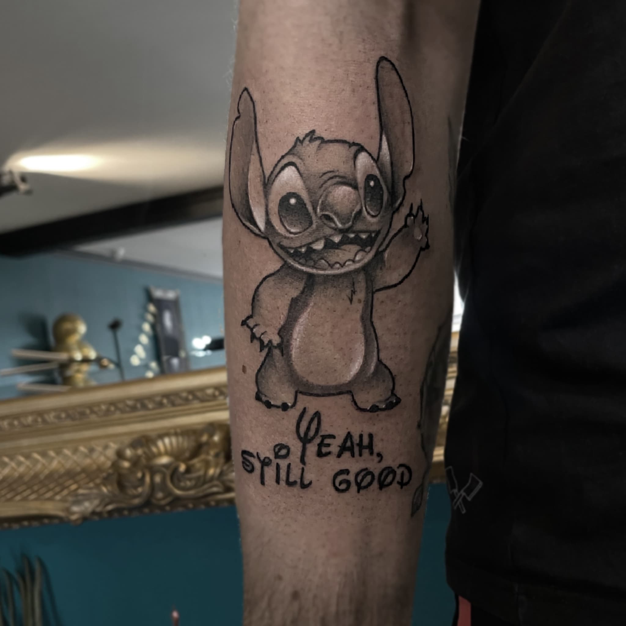 Zwart-wit tattoo van Stitch die zwaait met de tekst 'YEAH, STILL GOOD' eronder.