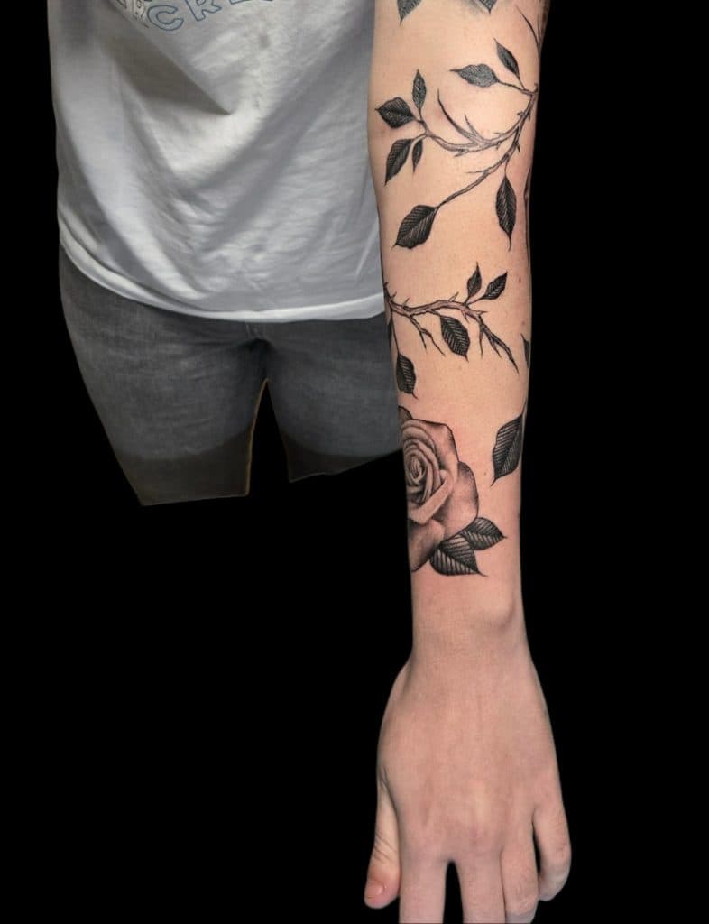 Zwart-wit tattoo van een roos met doornige takken en bladeren op de onderarm.