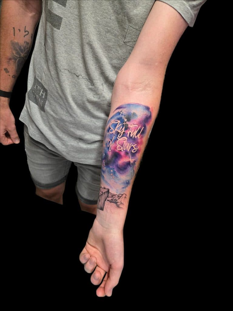 Watercolor tattoo met tekst 'Sky Full of Stars' en bergen op onderarm.