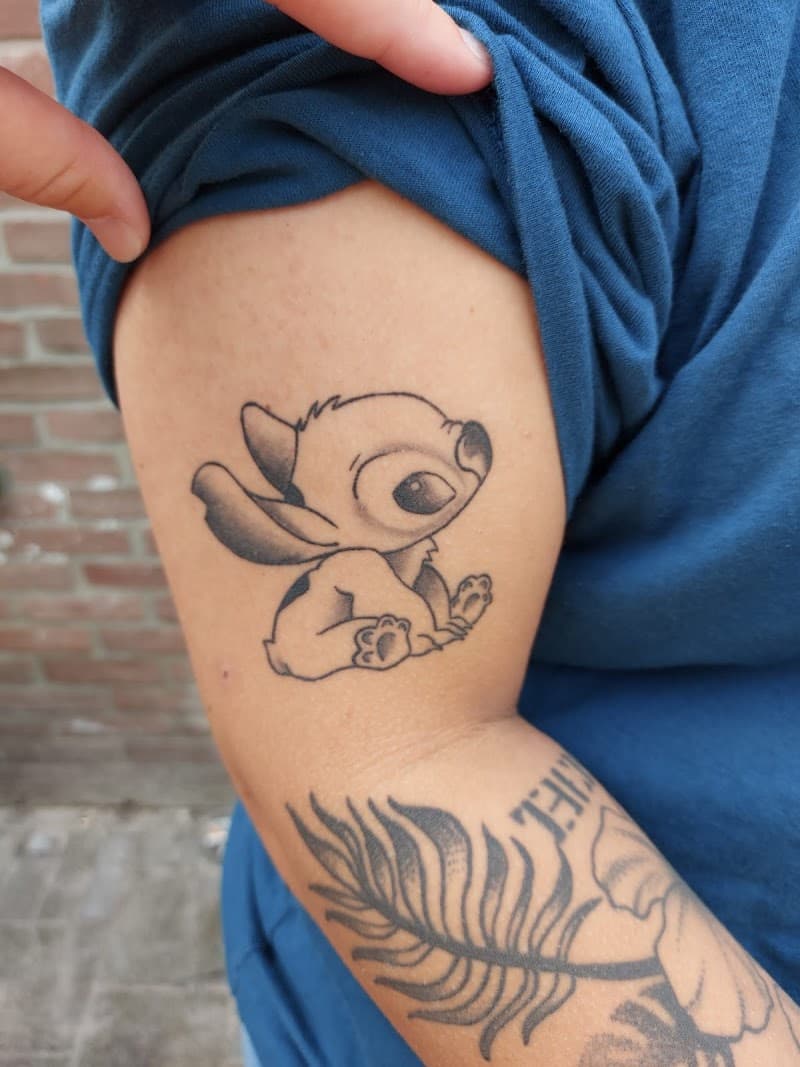 Schattige kleine Stitch tattoo in zwart-wit op de bovenarm.