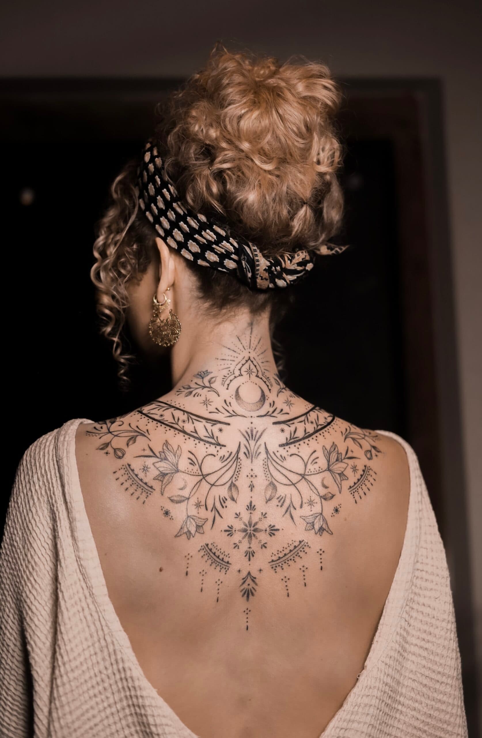 Grote fineline tattoo op nek en rug met maan, bloemen en ornamenten.