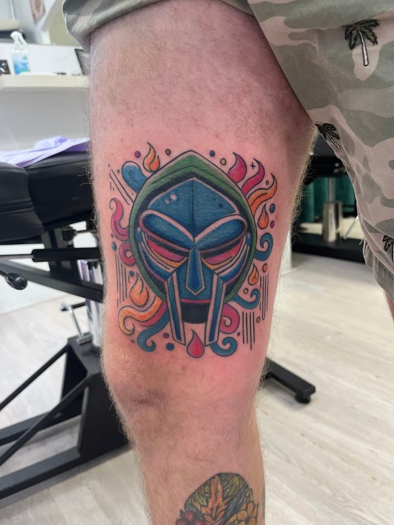 Kleurrijke illustratieve tattoo van het MF DOOM masker op het bovenbeen.