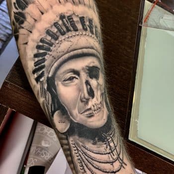 Realistische tattoo van een Indiaan met een half schedel gezicht op de onderarm.