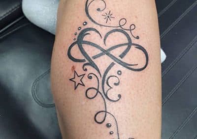 Blackwork tattoo van een infinity hart met krullen, sterren en stippen op onderbeen.
