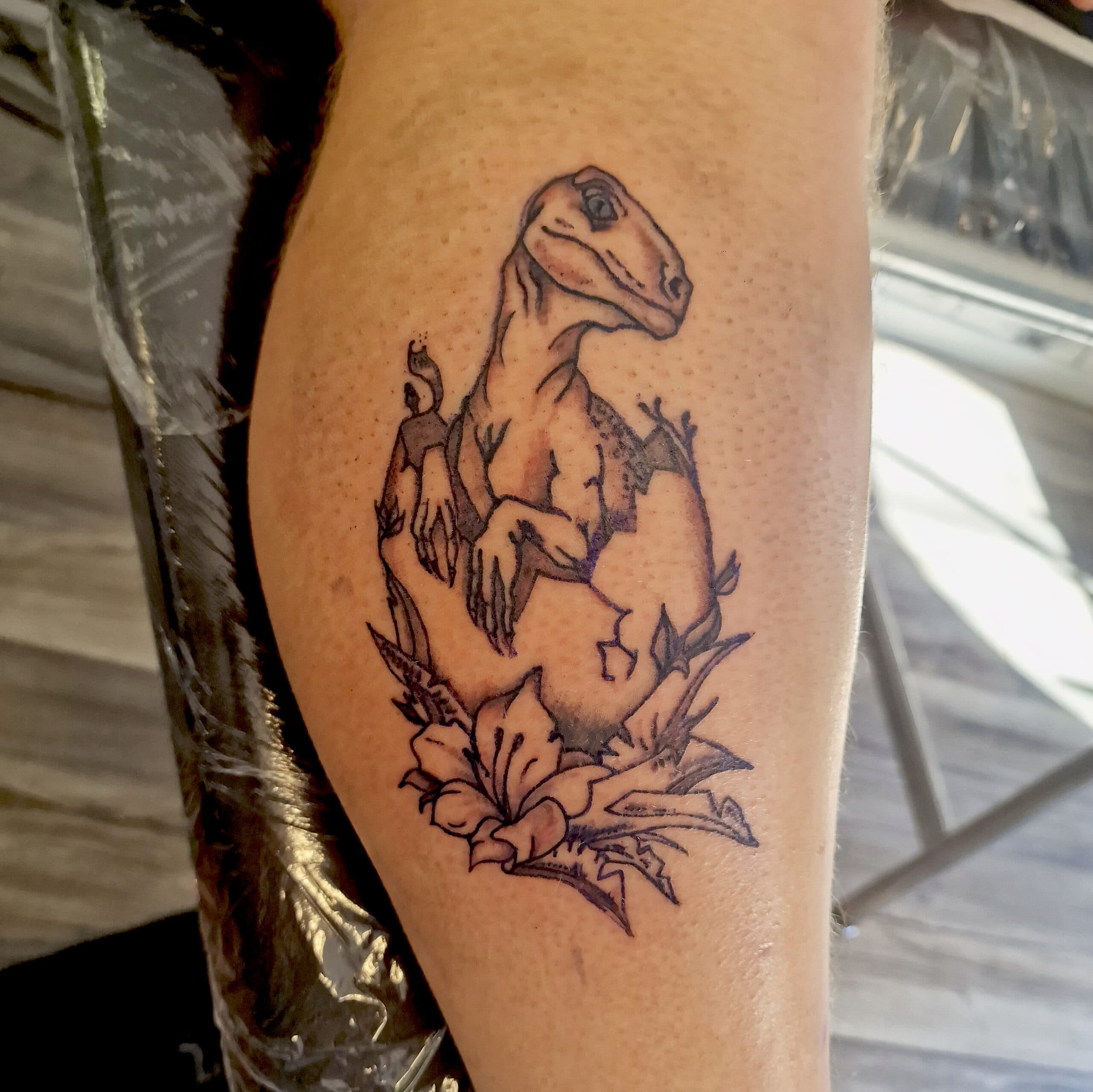 Illustratieve tattoo van een baby velociraptor die uit een ei komt met bloemen.
