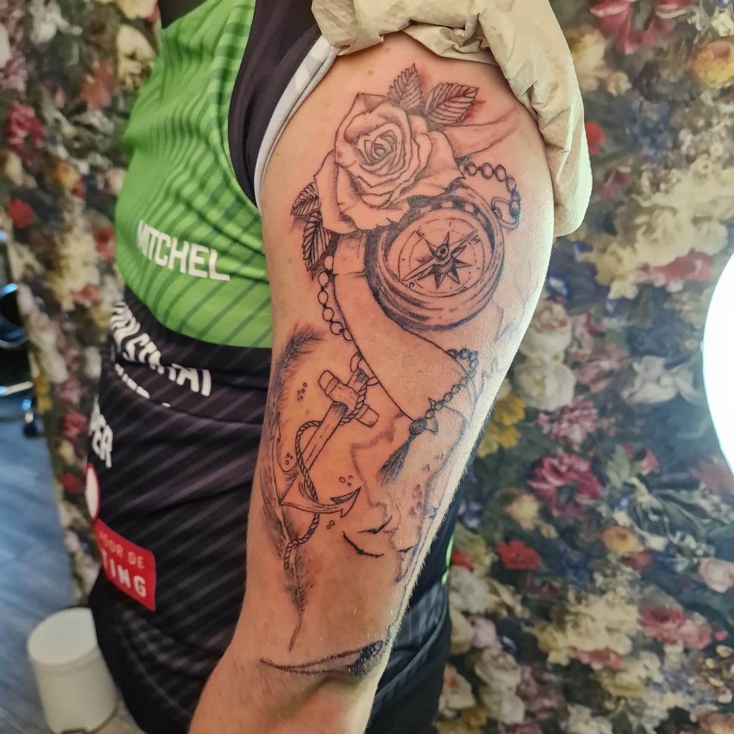 Zwart-wit tattoo van een roos, kompas, anker en veer op de bovenarm.