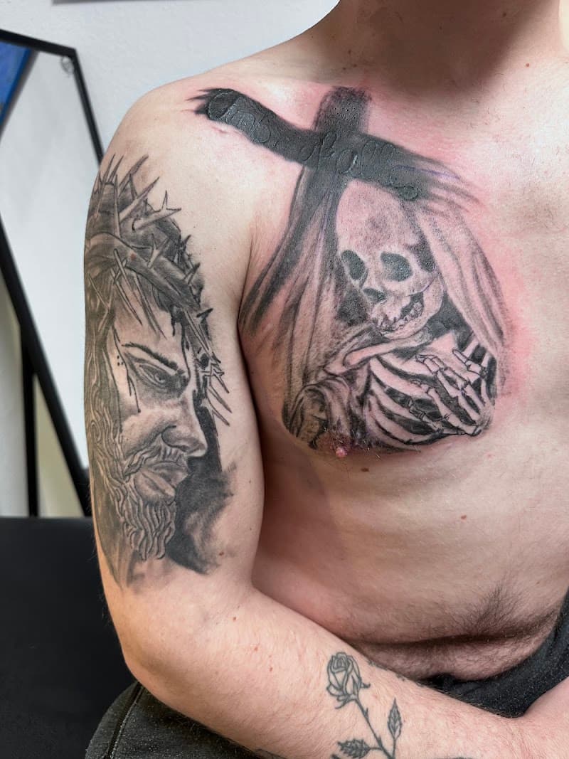 Zwart-wit tattoos van Jezus, skelet met kruis en roos op arm en borst.