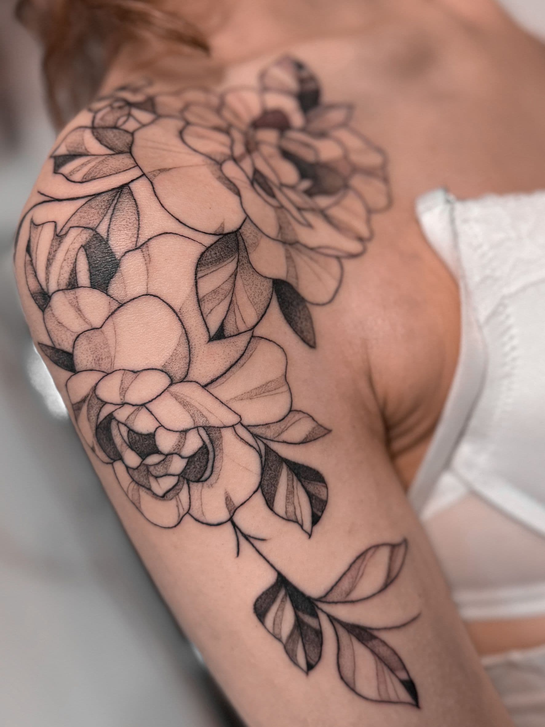 Elegante zwart-wit bloemen tattoo op schouder, fineline en dotwork stijl.