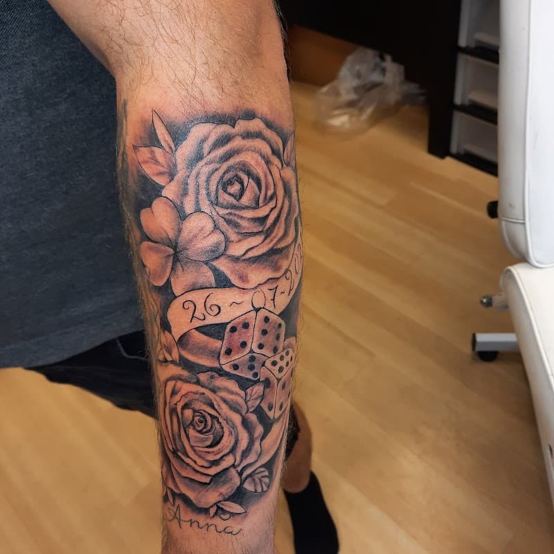 Zwart-wit tattoo van rozen, dobbelstenen, datum en naam 'Anna' op onderarm.