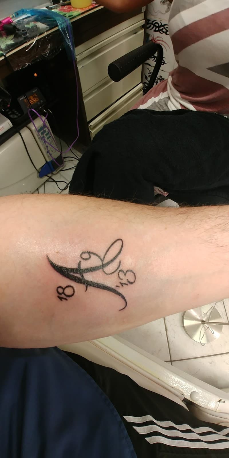 Zwart-wit tattoo van een sierlijke letter 'A' met de getallen '18' en '13'.