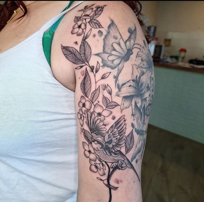 Fineline vogel en bloemen tattoo op bovenarm, met oudere tattoo erachter.