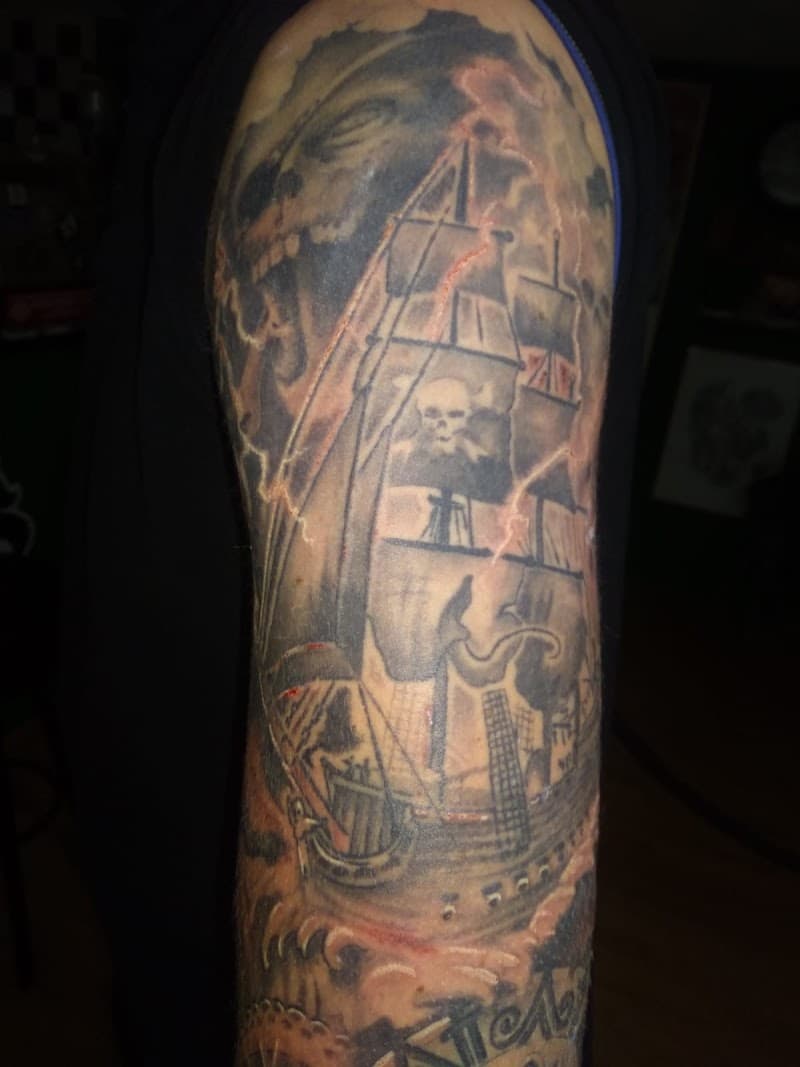 Zwart-wit tattoo van een piratenschip en een schedel in de wolken op de bovenarm.