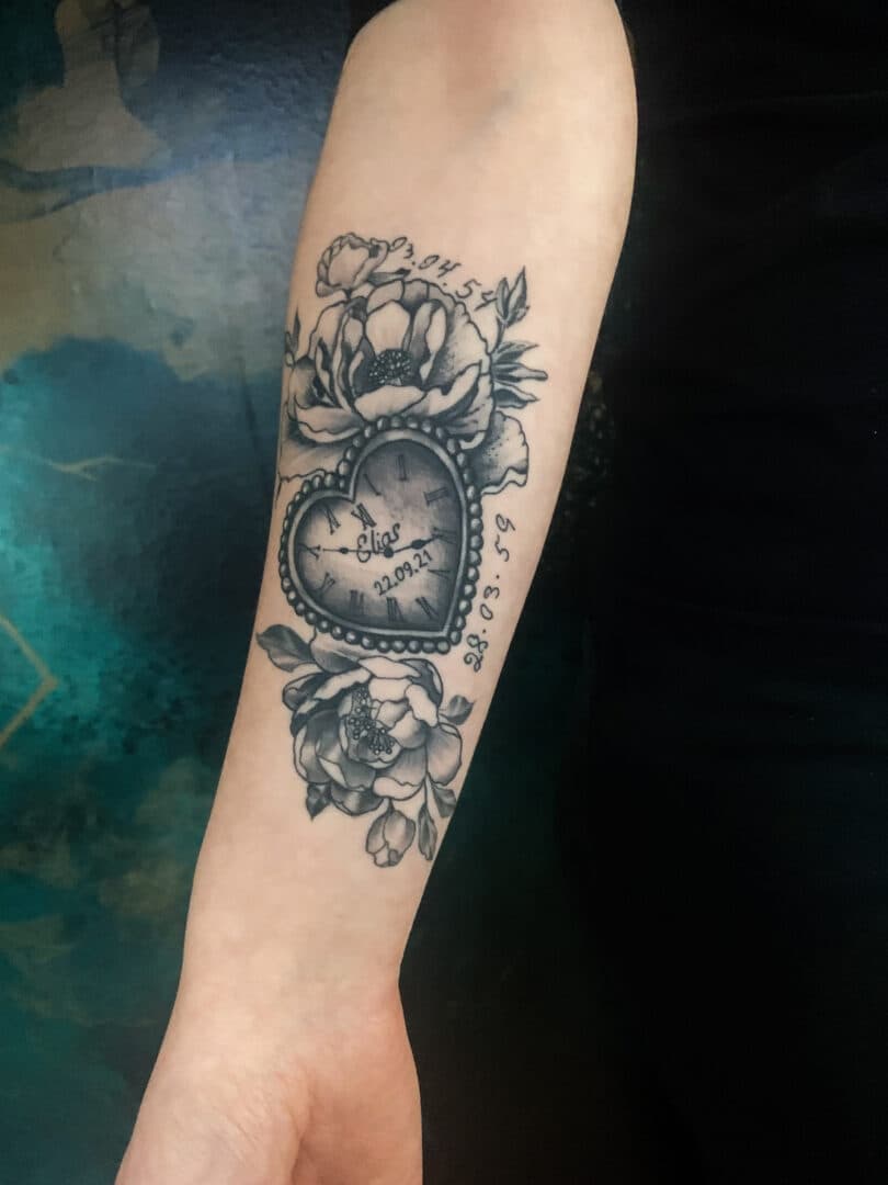 Blackwork tattoo van een hartvormige klok met bloemen en datums op de onderarm.