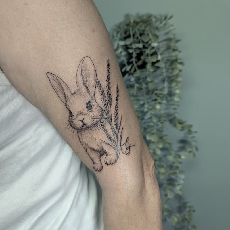 Fineline tattoo van een konijn met graanhalmen op de bovenarm.