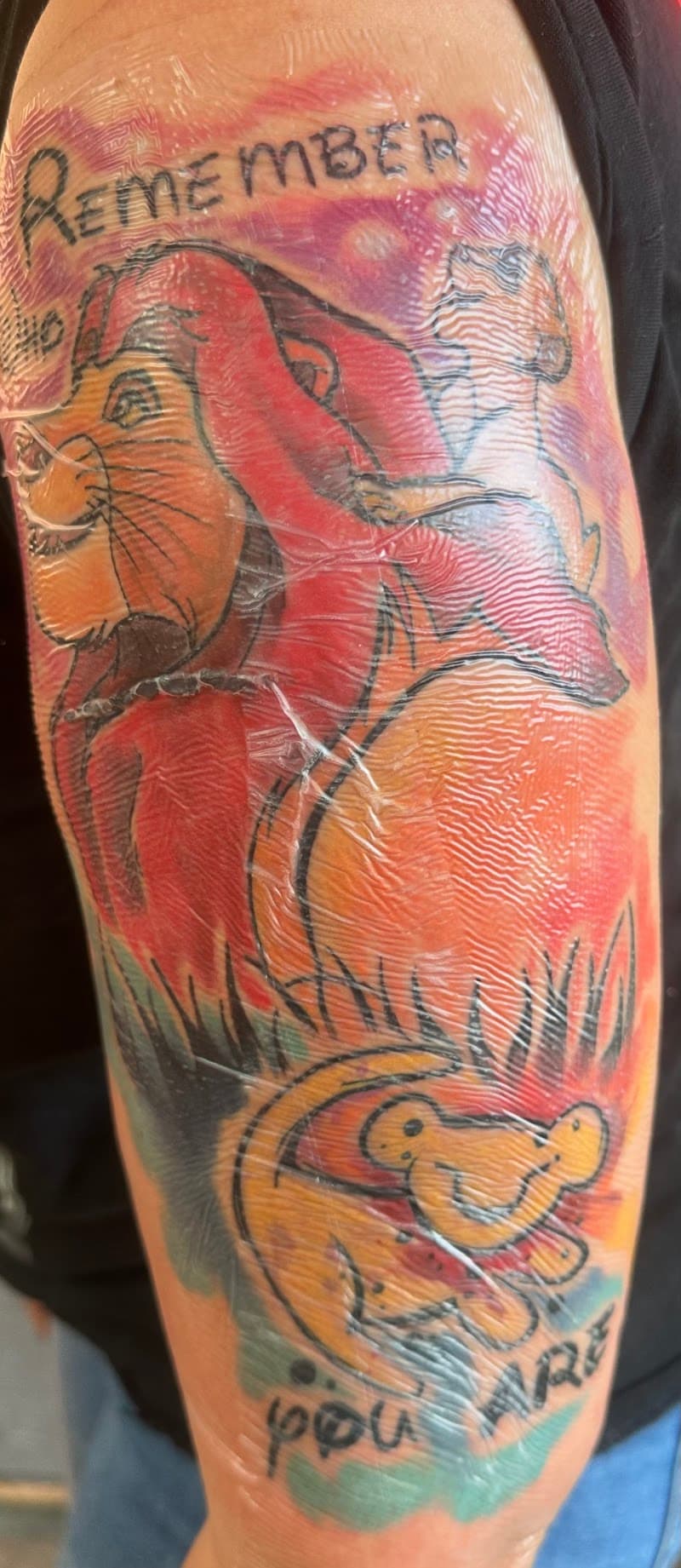 Kleurrijke Lion King tattoo met Mufasa, Simba, Timon, Pumbaa en tekst.