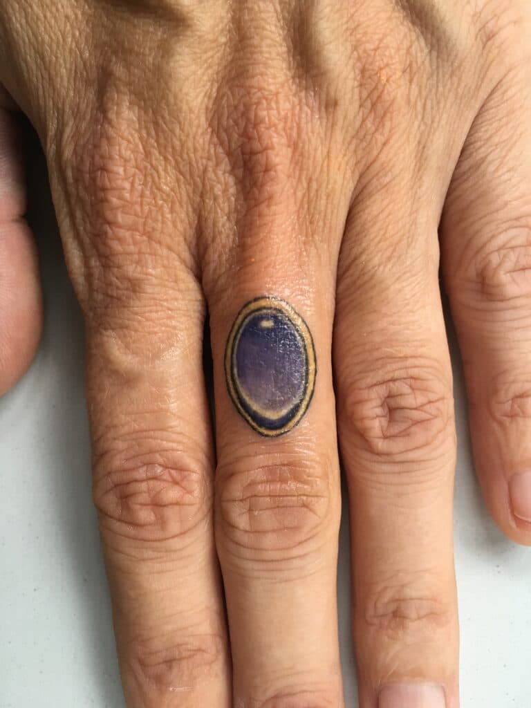 Kleine ovale edelsteen ring tattoo op de ringvinger.
