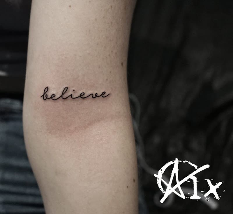 Minimalistische 'believe' tekst tattoo in sierlijk handschrift op de bovenarm.