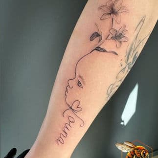 Fineline tattoo van baby profiel, naam 'Laura' en lelies op onderarm.