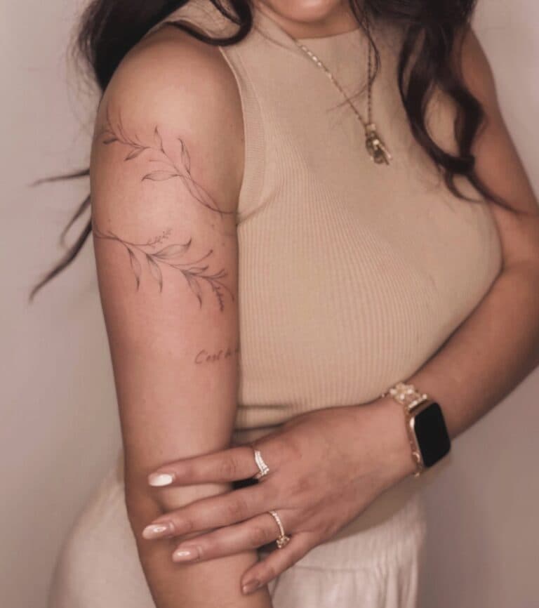 Fineline tak met bladeren en kleine tekst op bovenarm.