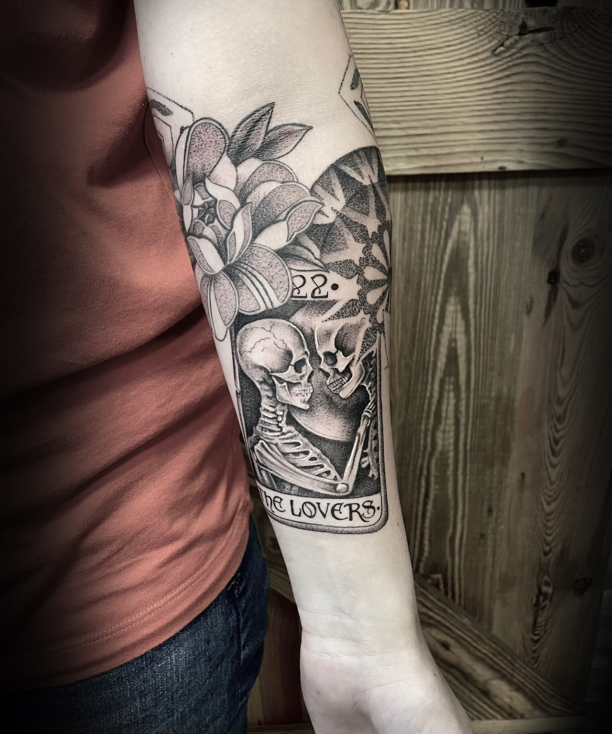 Blackwork tarotkaart 'The Lovers' met skeletten en bloemen op bovenarm.