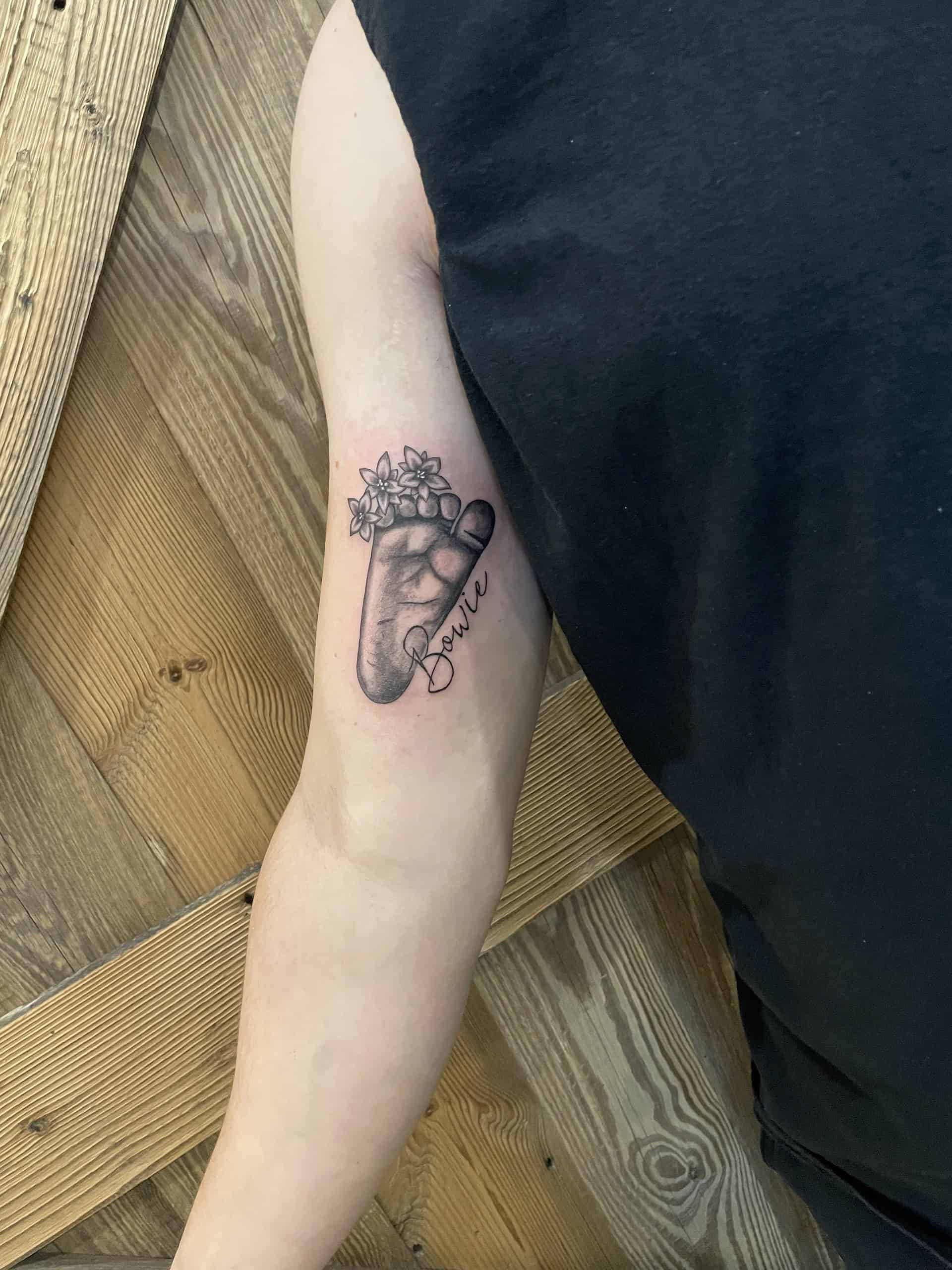 Realistische baby voetafdruk met bloemen en naam 'Bowie' op bovenarm.