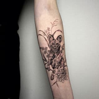 Fineline tattoo van een wilde hond met bloemen op de onderarm.