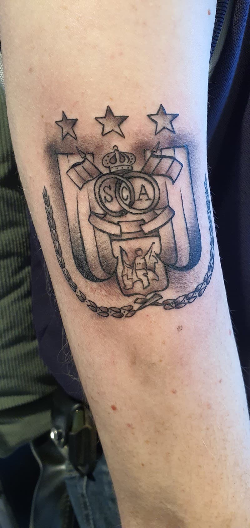 Zwart-wit wapenschild tattoo met 'SCA', kroon, sterren en vlaggen op bovenarm.