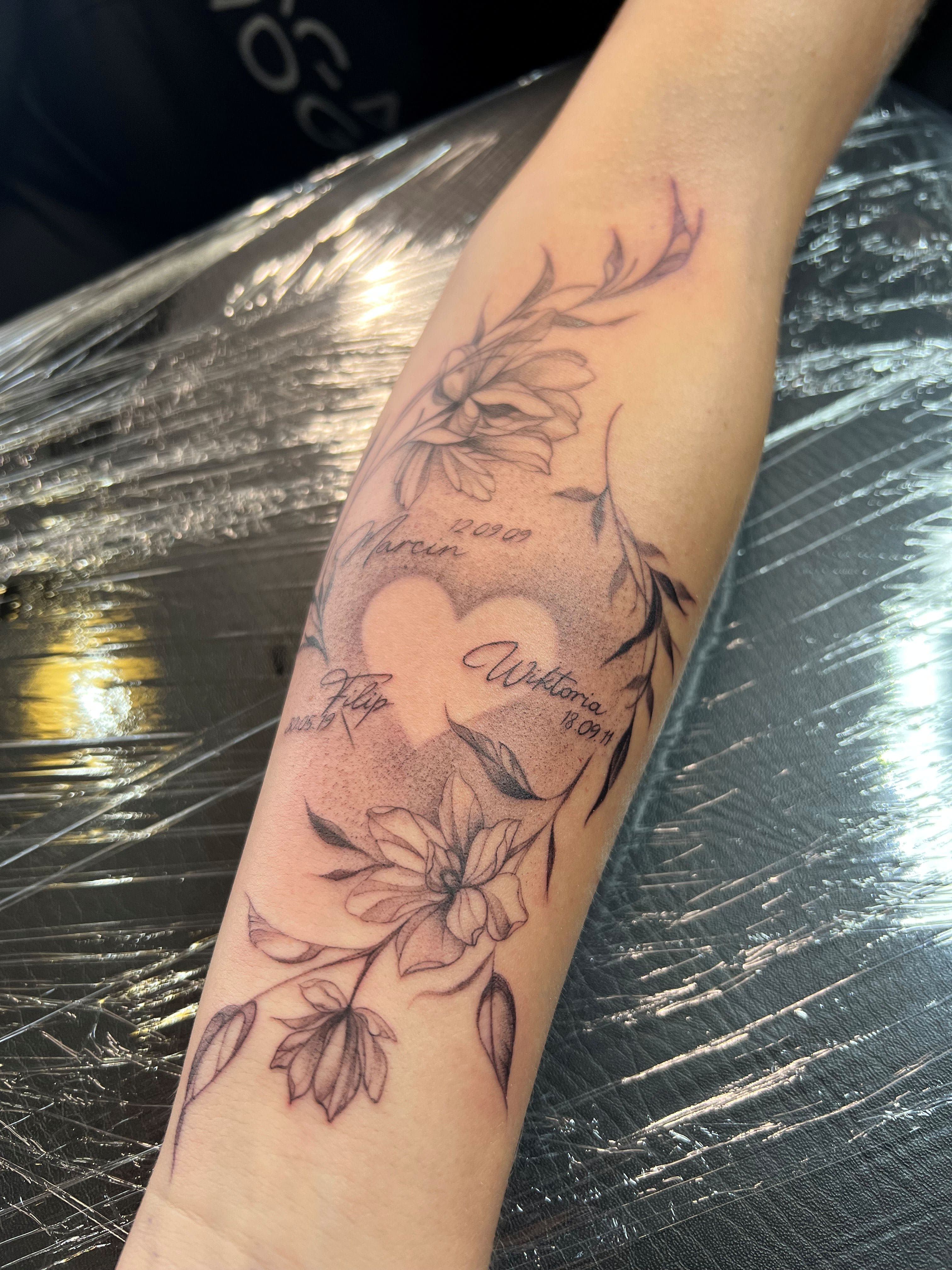 Fineline bloemen en hart met namen en datums op onderarm.