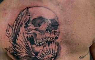 Zwart-wit tattoo van een schedel met vleugels en een duif op de borst.