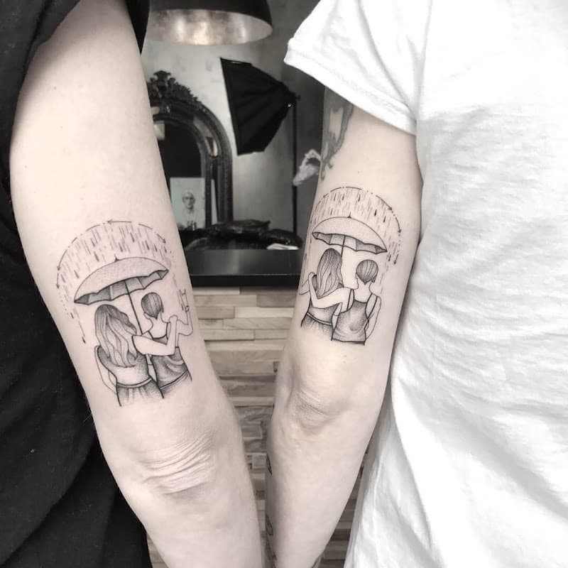Fineline tattoo van twee figuren onder een paraplu in de regen op twee bovenarmen.