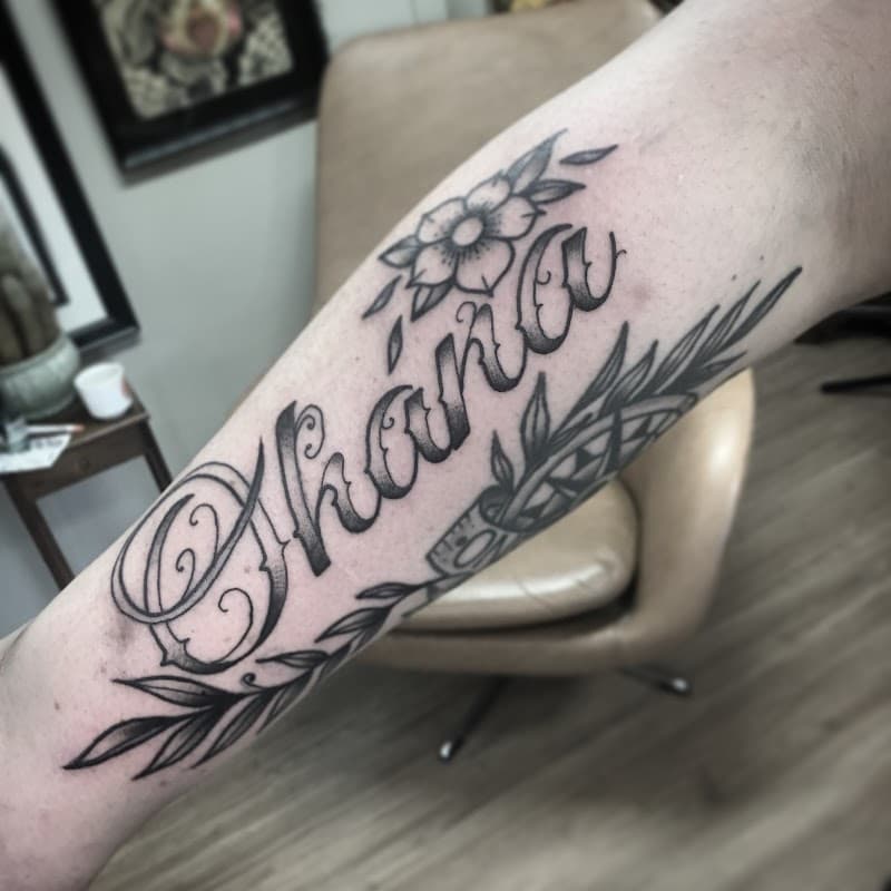 Zwart-wit 'Ohana' tekst met bloemen en bladeren op onderarm.