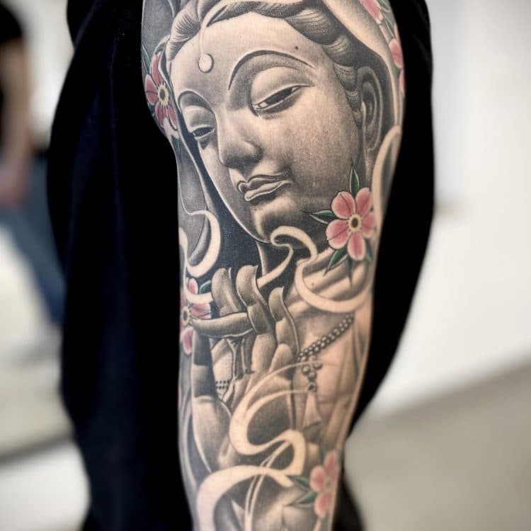 Black & grey tattoo van Boeddha/Guanyin met roze bloemen op de bovenarm.