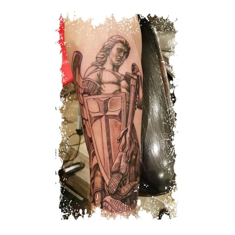 Zwart-wit tattoo van Aartsengel Michaël met schild en zwaard op de bovenarm.