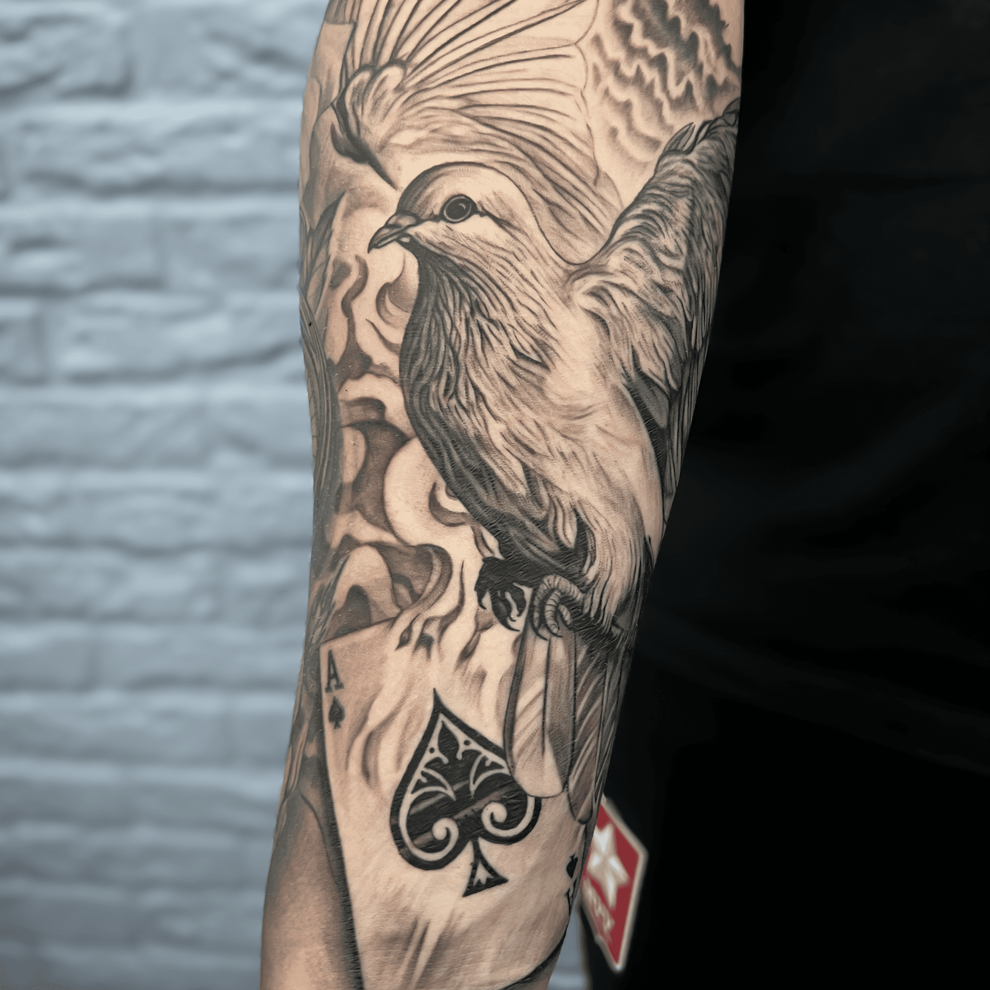 Zwart-wit tattoo van een duif en een schoppenaas kaart op de bovenarm.