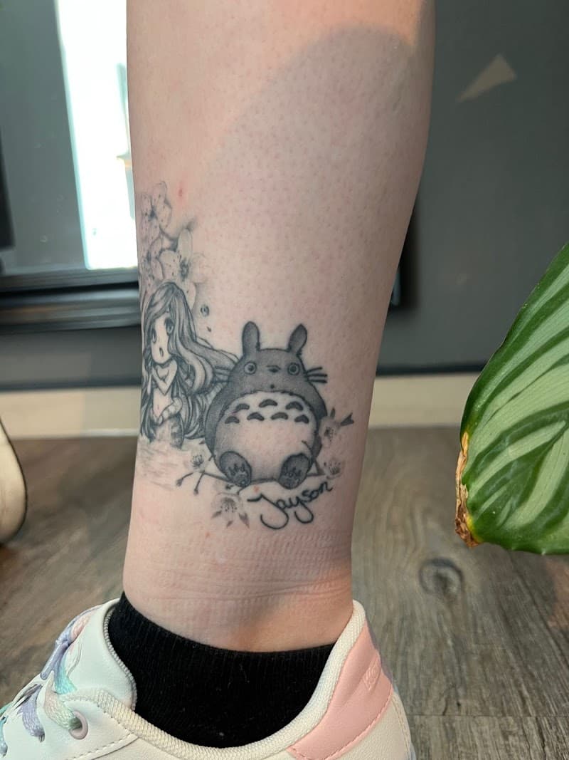 Zwart-wit tattoo van Totoro en een meisje met bloesem op de enkel.