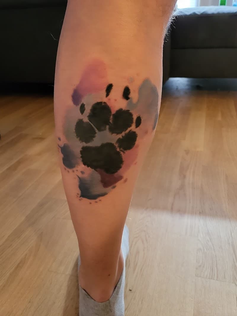 Aquarel hondenpoot tattoo met roze, paarse en blauwe spetters op onderbeen.
