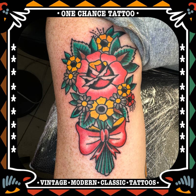 Traditionele tattoo van een roos met bloemen en een strik op het been.