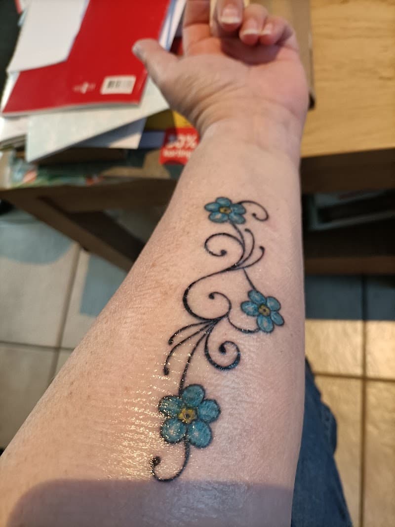 Sierlijke tattoo van drie blauwe bloemen met zwarte krullen op de onderarm.