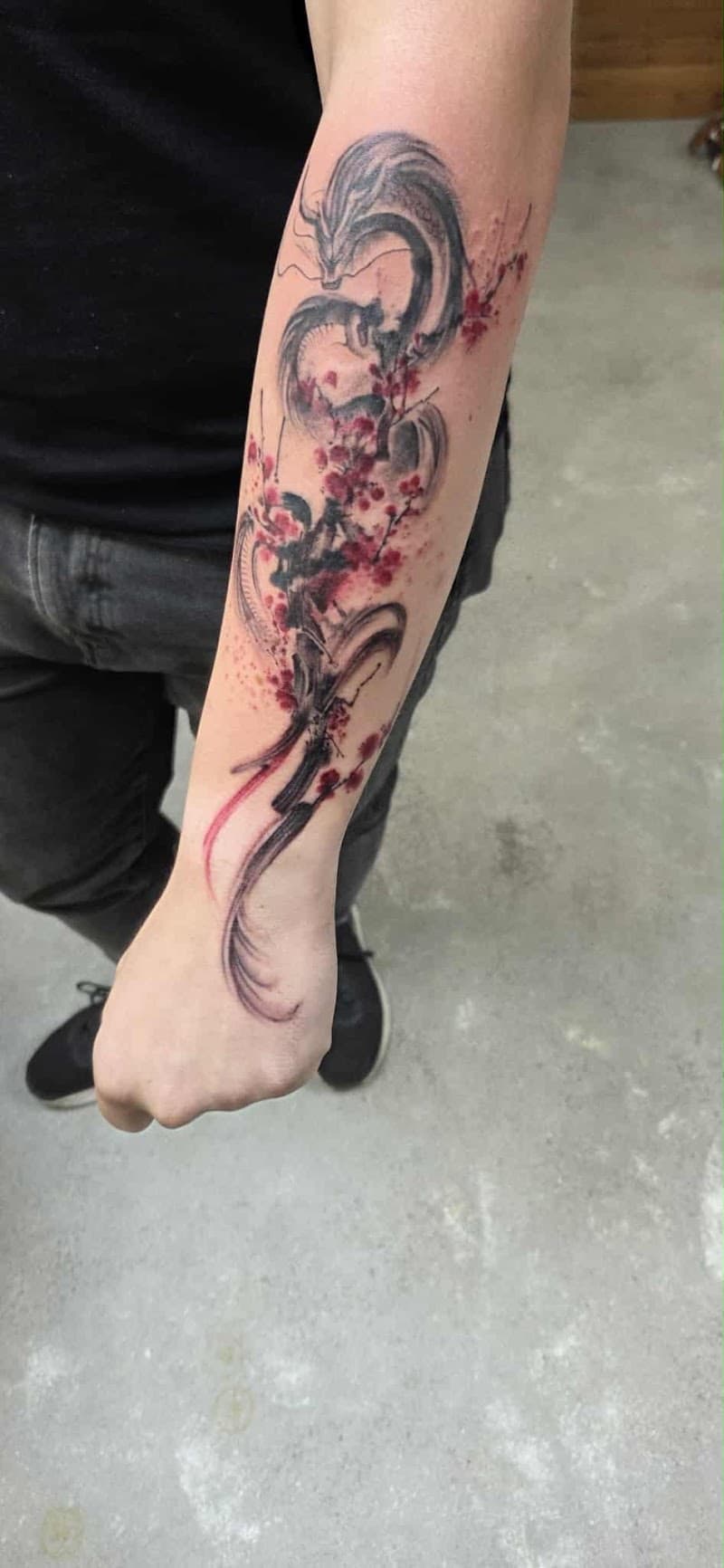 Zwart-grijze draak met rode kersenbloesem in watercolor stijl op onderarm.