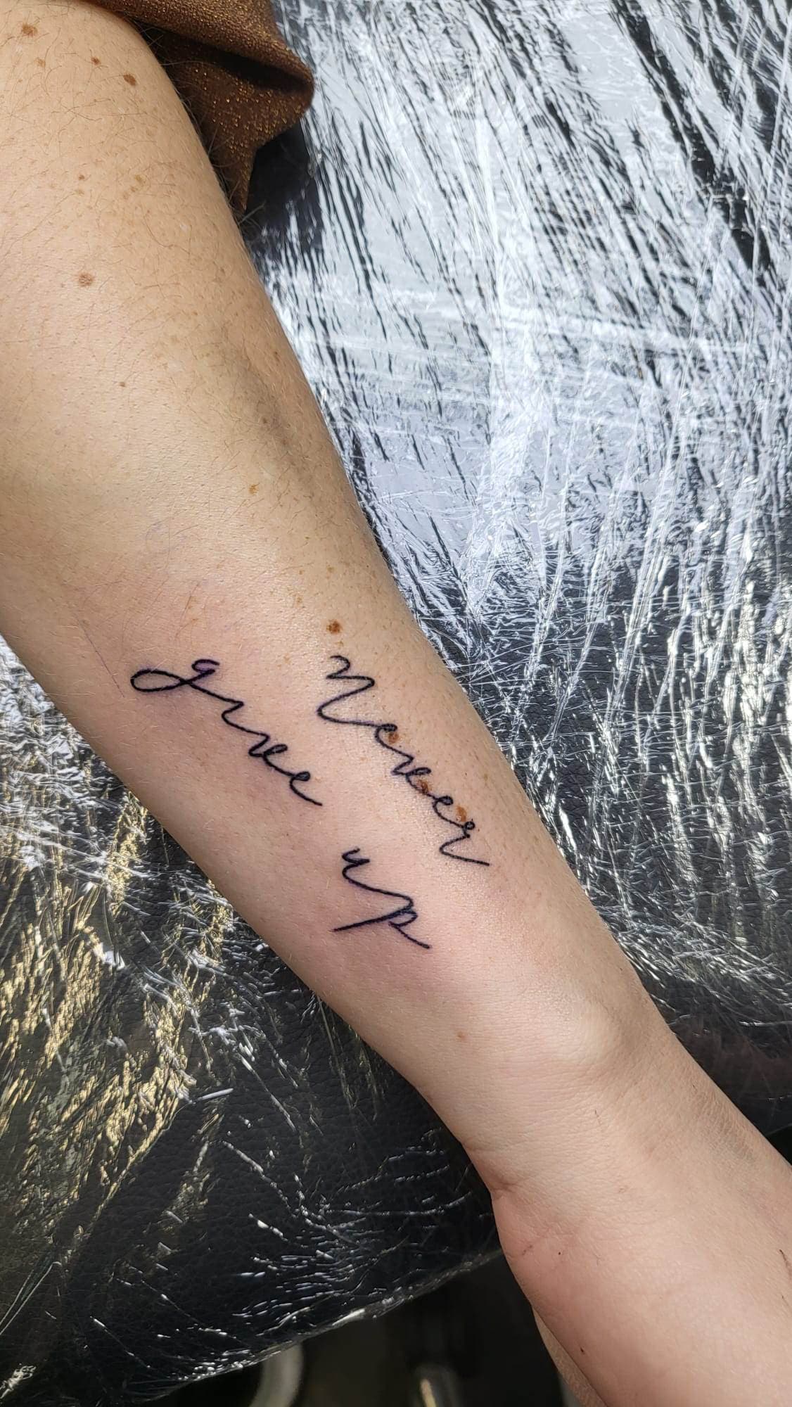 "Never give up" tekst tattoo in sierlijk handschrift op onderarm.