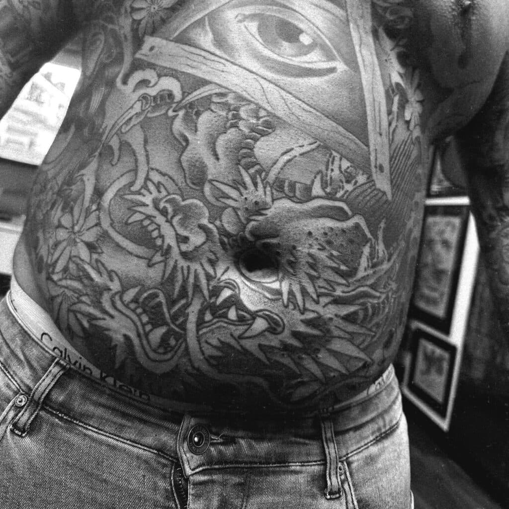 Zwart-wit tattoo op de buik met een draak, een alziend oog in een driehoek en bloemen.