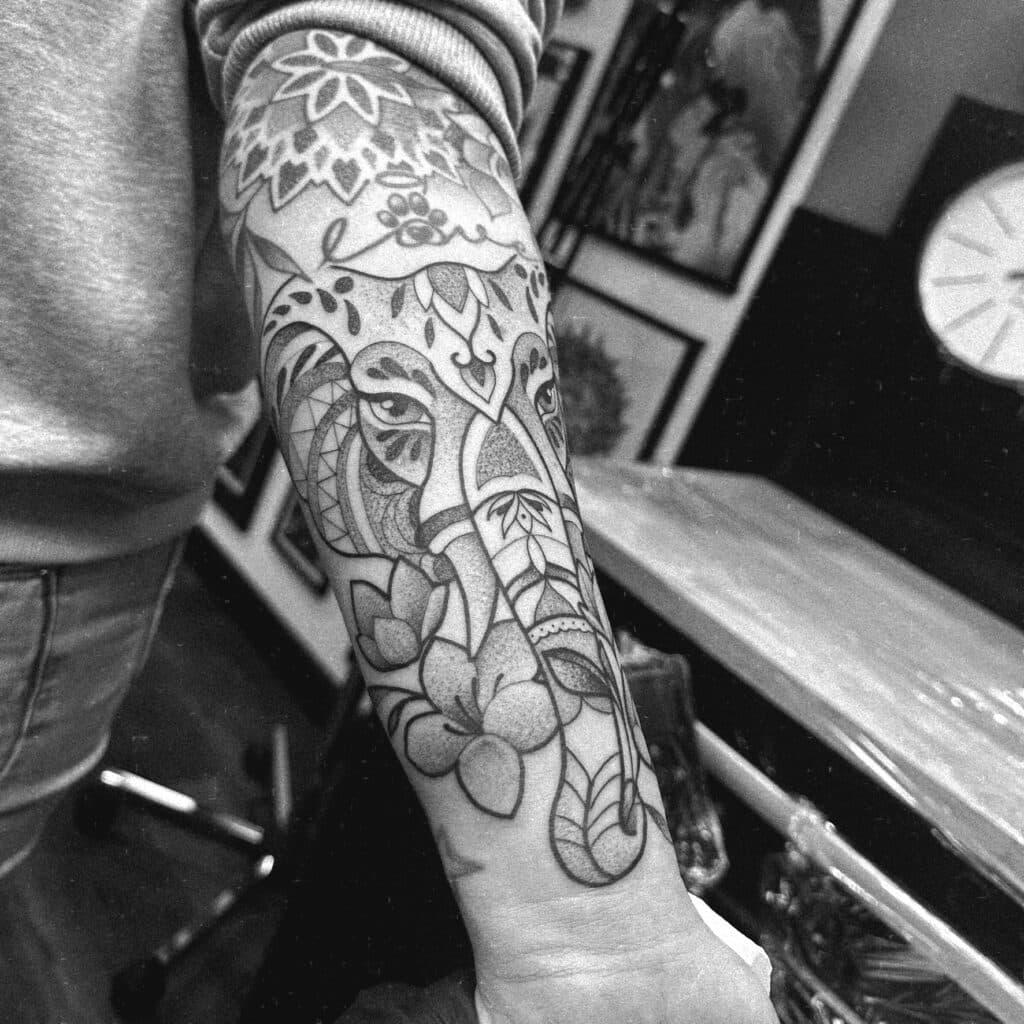 Zwart-wit olifant tattoo met mandala en bloemen op onderarm.