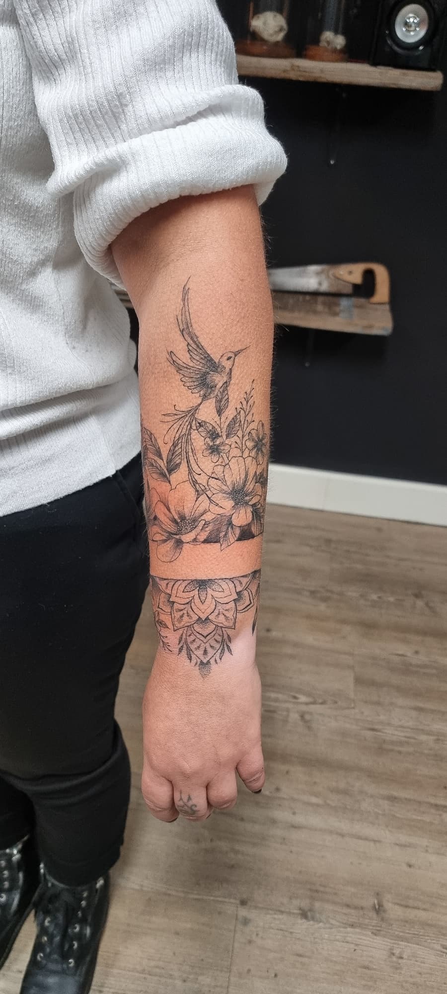 Fineline tattoo van een kolibrie met bloemen en een mandala band op de onderarm.