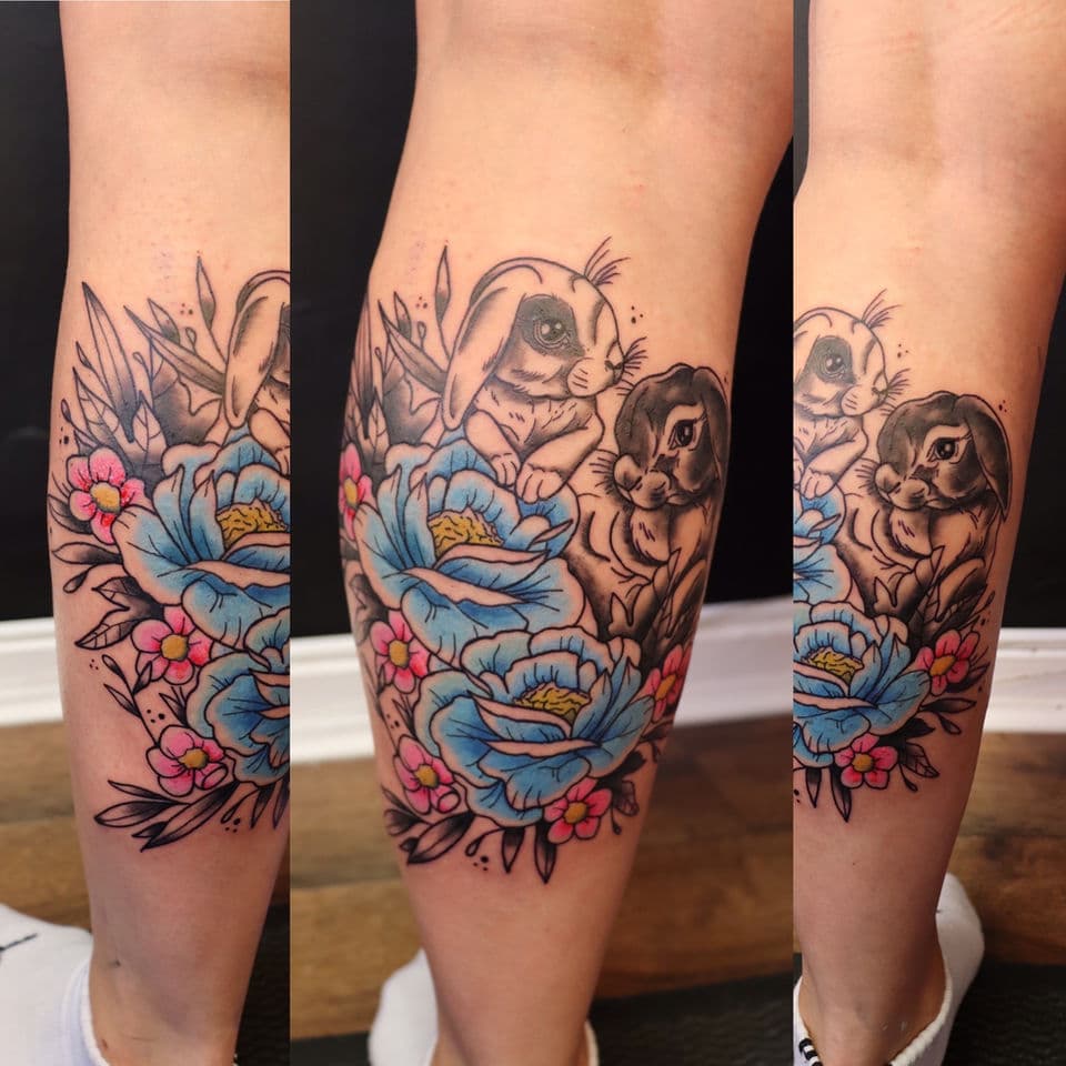 Neo-traditionele tattoo van twee konijnen met blauwe en roze bloemen op onderbeen.
